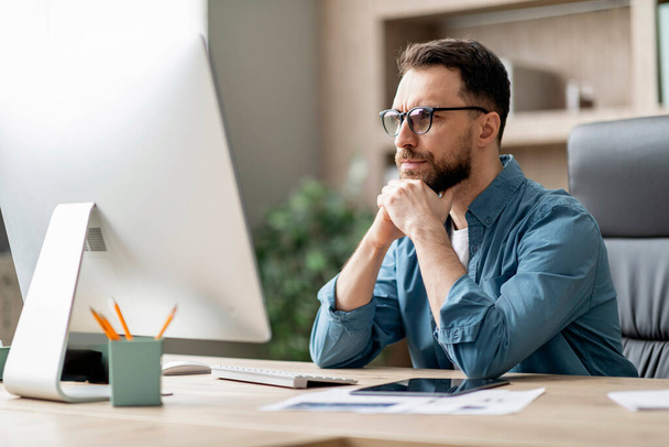 Портрет застенчивого молодого бизнесмена в очках, смотрящего на экран компьютера во время работы за столом в современном офисе, фокусирующегося на мыслях предпринимателя тысячелетия о бизнес-стратегии - Фото, изображение