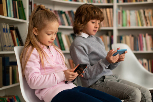 Okul çocukları tatilden sonra akıllı telefon kullanıyor, oyun oynayan çocuklar veya cep telefonlarıyla video seyrediyor, sandalyeye oturuyorlar. - Fotoğraf, Görsel