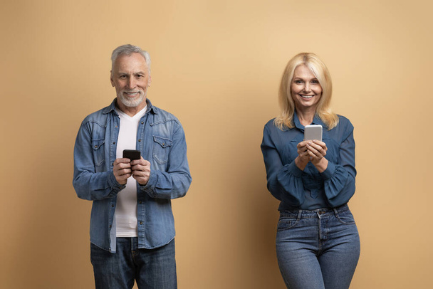 Szczęśliwy starszy mężczyzna i kobieta ubrani w dżinsy, używający telefonów komórkowych na beżowym tle studia. Emerytowani starsi ludzie przewijanie, websurfing, za pomocą randkowej aplikacji mobilnej - Zdjęcie, obraz