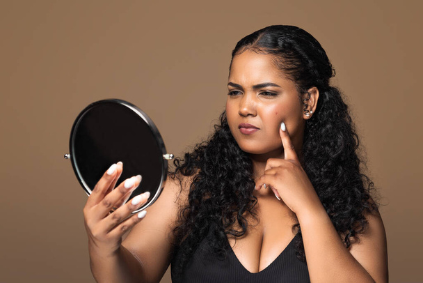 Skin disorders. Unhappy sad brazilian body positive woman looking at mirror, lady touching pimples on her cheek skin, standing over brown studio background - Foto, Bild