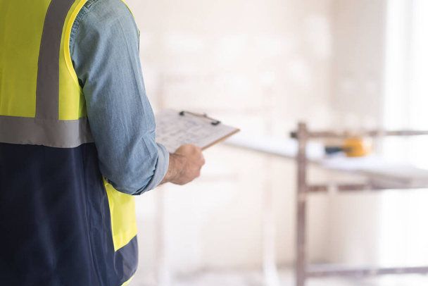 Профессиональный инженер держит планшет с планом ремонта интерьера дома в руках, стоящих напротив обветшалой стены и строительной техники в светлом помещении - Фото, изображение