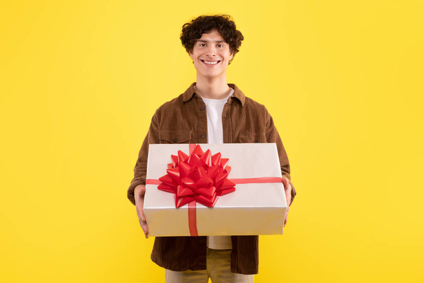 Senin için en iyi hediye. Sarılı hediye kutusunu tutan adamın ön manzara portresi, kameraya gülümsüyor, sarı stüdyo arka planında poz veriyor. Özel bayram gününü kutlamak ve selamlamak - Fotoğraf, Görsel