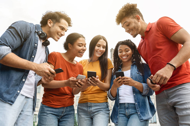Tolle App. Gruppe multiethnischer Studenten, die gemeinsam Smartphones benutzen, während sie sich im Freien entspannen, glückliche junge Leute, die auf den Handybildschirm schauen, neue Studie App oder Website durchsuchen, Nahaufnahme - Foto, Bild