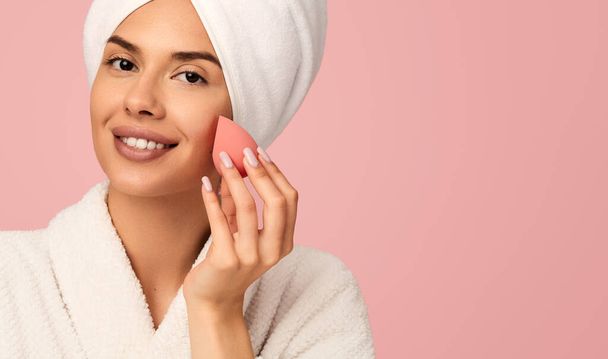 Positive young female in bathrobe and towel on head applying makeup with pink sponge on face, while against pink background with copy space - 写真・画像