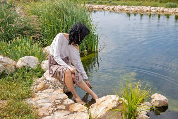 Mooie jonge brunette Kaukasische vrouw zitten in de buurt van het meer raakt het water en kijkt naar het close-up. - Foto, afbeelding