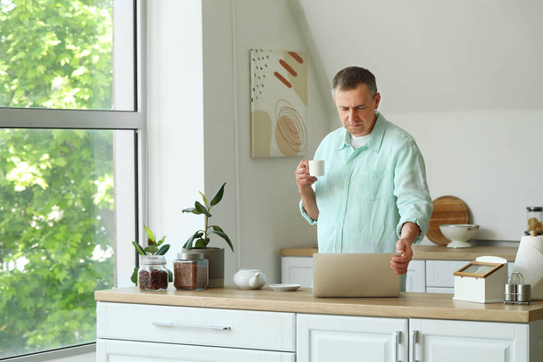 Dojrzały mężczyzna z filiżanką kawy i laptop wiadomości w kuchni - Zdjęcie, obraz
