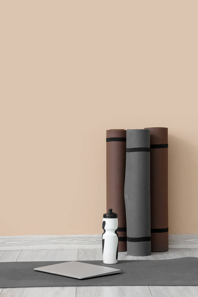 Фітнес килимки з пляшкою води і ноутбуком біля бежевої стіни в кімнаті - Фото, зображення