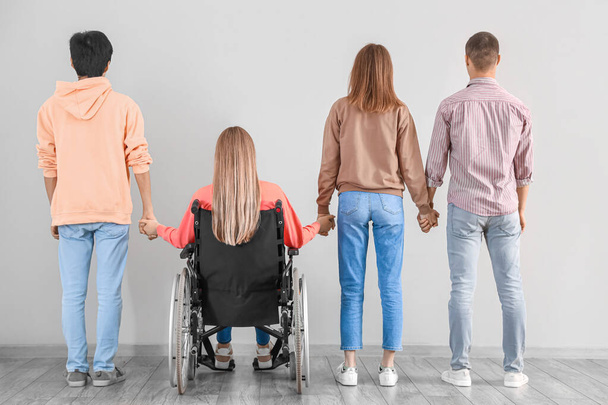 Группа подростков с девушкой в инвалидной коляске держатся за руки возле светлой стены, вид сзади - Фото, изображение