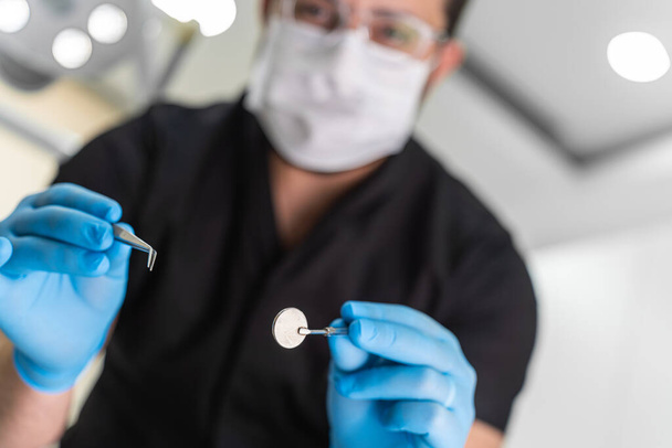 Dentista masculino experto en uniforme negro que examina la cavidad oral del paciente con pinzas metálicas y espejo redondo en la cita en el centro de estomatología procedimiento de cuidado de dientes - Foto, imagen