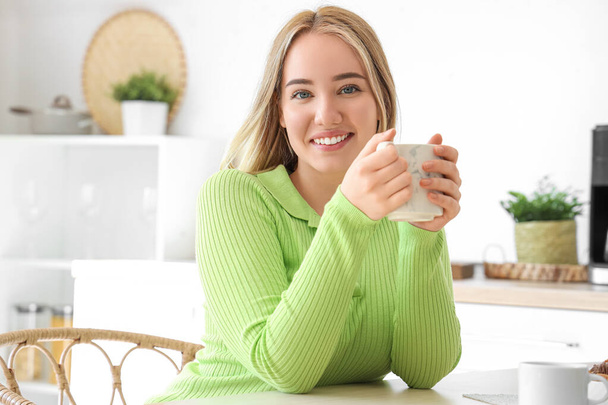 Młoda kobieta z filiżanką gorącej herbaty siedzi przy stole w kuchni - Zdjęcie, obraz