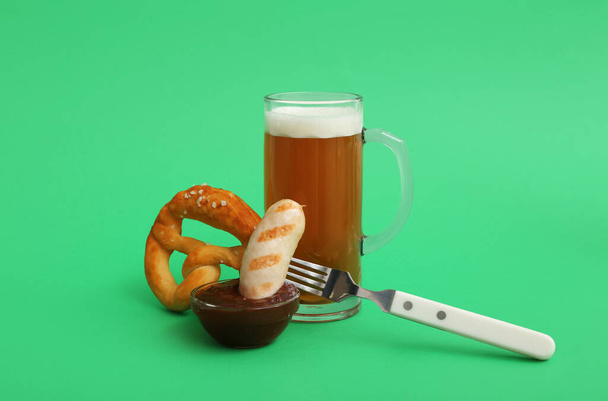 Widelec ze smaczną bawarską kiełbasą, precelkiem, sosem i kubkiem piwa na zielonym tle. Święto Oktoberfest - Zdjęcie, obraz
