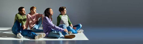 νέοι άνδρες σε casual φορούν κάθεται στο πάτωμα κοιτάζοντας μακριά σε γκρι φόντο, πολιτιστική πολυμορφία, πανό - Φωτογραφία, εικόνα