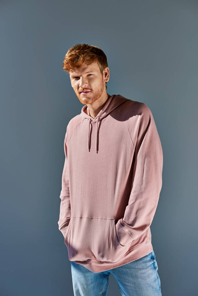 stylish young red haired man in pink hoodie and jeans looking at camera posing on grey backdrop - Foto, imagen