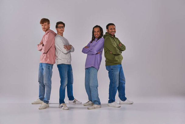 four young men standing back to back and smiling at camera on grey backdrop, cultural diversity - Foto, afbeelding