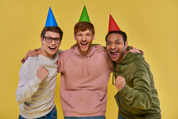 three multicultural cheerful friends in birthday hats hugging and looking at camera, birthday - Photo, Image