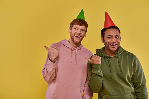 dos jóvenes amigos en atuendo casual y sombreros de cumpleaños sonriendo a la cámara señalando los pulgares a un lado - Foto, Imagen