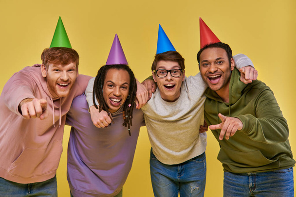 amigos multiculturales en sombreros de cumpleaños abrazándose mirando y sonriendo a la cámara, cumpleaños - Foto, imagen