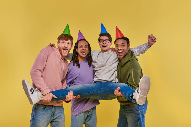 czterech wesołych mężczyzn w urodzinowych kapeluszach uśmiechniętych do kamery i trzymających jednego z nich na rękach, urodziny - Zdjęcie, obraz