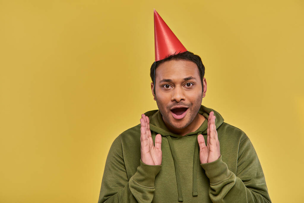 astonished young indian man in birthday hat and khaki hoodie with hands closer to face, birthday - Fotoğraf, Görsel