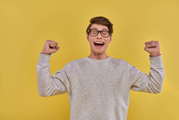 эмоциональный счастливый человек в повседневной уличной одежде поднимает кулаки вверх и закрывает глаза на жёлтом фоне - Фото, изображение