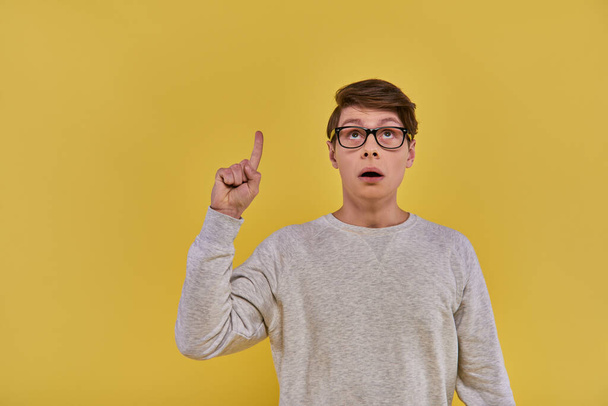 young man in white sweatshirt raising his finger in eureka gesture and looking up on yellow backdrop - Photo, Image