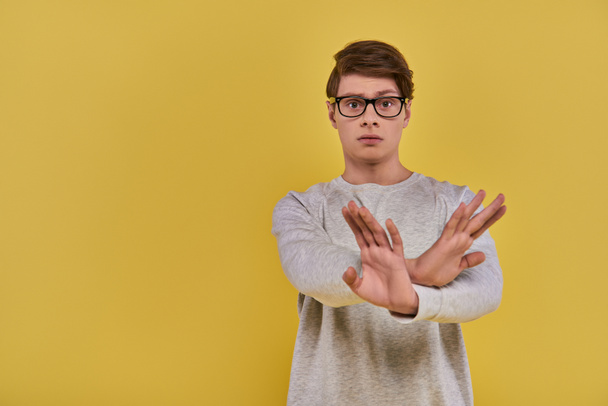 young sad man in white sweatshirt with glasses showing denial cross hand gesture on yellow backdrop - Photo, Image