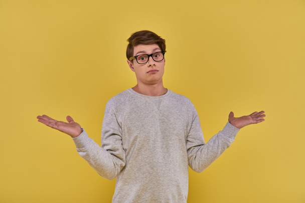 смущенный невежественный молодой человек в толстовке и очках показывает беспомощный жест на желтом фоне - Фото, изображение