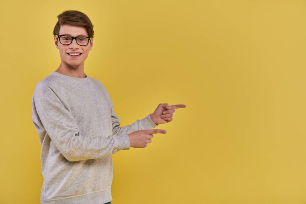 joyeux jeune homme en tenue décontractée pointant deux doigts de côté sur le côté droit sur fond jaune - Photo, image