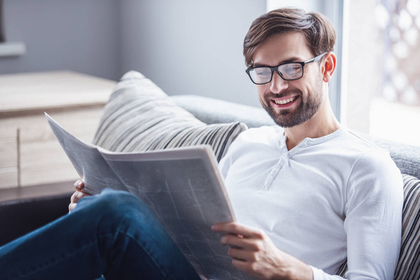 Handsome man in eyeglasses is reading a newspaper and smiling while sitting on couch at home - Photo, Image
