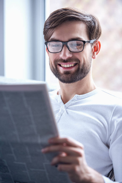Hombre guapo con anteojos está leyendo un periódico y sonriendo mientras está sentado en el sofá en casa
 - Foto, imagen