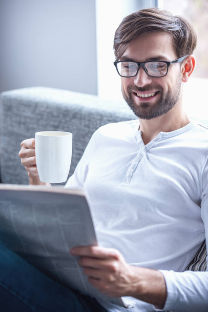 Handsome man in eyeglasses is reading a newspaper, holding a cup and smiling while sitting on couch at home - Valokuva, kuva