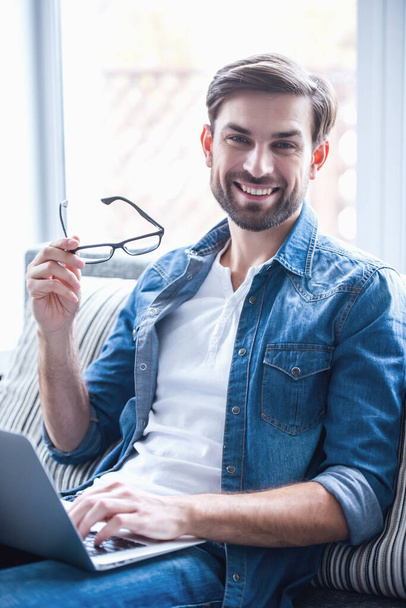 Hombre de negocios guapo está sosteniendo gafas, mirando a la cámara y sonriendo mientras trabaja con un ordenador portátil en casa
 - Foto, Imagen