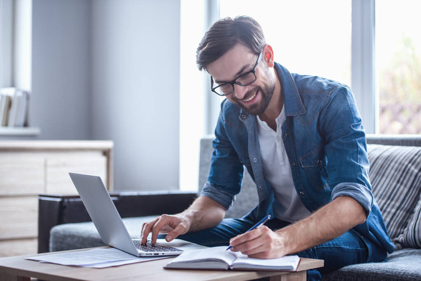 Όμορφος επιχειρηματία σε γυαλιά κάνει σημειώσεις και χαμογελώντας ενώ εργάζεστε με ένα φορητό υπολογιστή στο σπίτι - Φωτογραφία, εικόνα