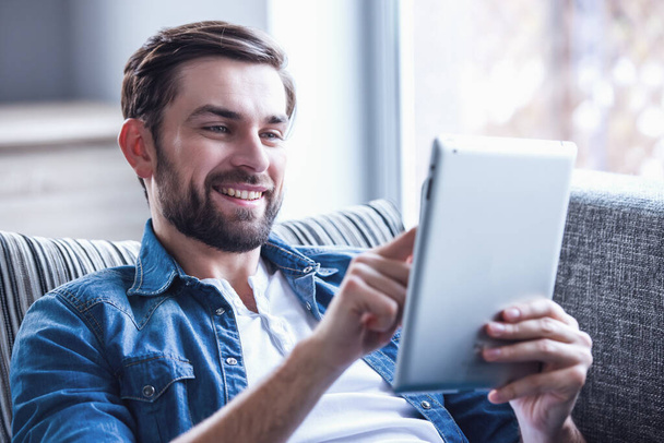 Yakışıklı adam dijital tablet kullanıyor ve evinde kanepede dinlenirken gülümsüyor. - Fotoğraf, Görsel