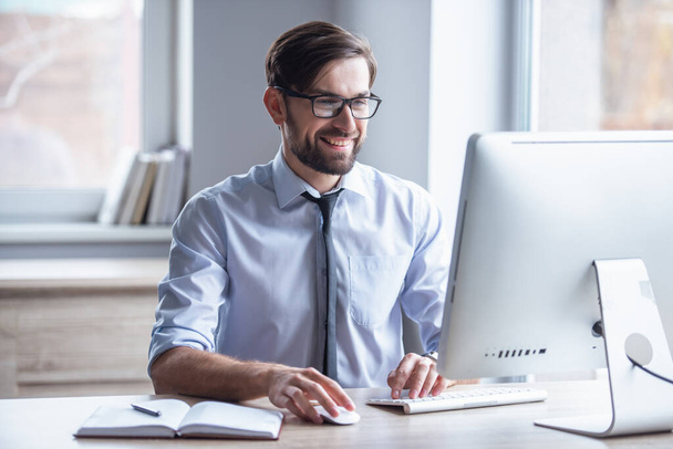 Bello uomo d'affari in occhiali sta usando un computer e sorridente mentre lavora in ufficio
 - Foto, immagini