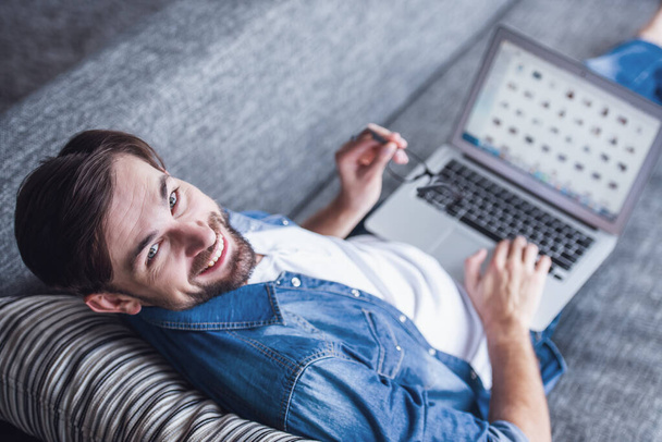 Jóképű férfi használ egy laptop, kamera nézett és mosolyogva, miközben feküdt a kanapén, otthon - Fotó, kép
