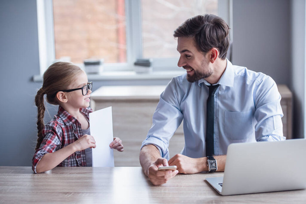 gutaussehender Geschäftsmann benutzt einen Laptop, während er zu Hause arbeitet. seine süße kleine Tochter in Brille zeigt ihm ein Blatt Papier - Foto, Bild