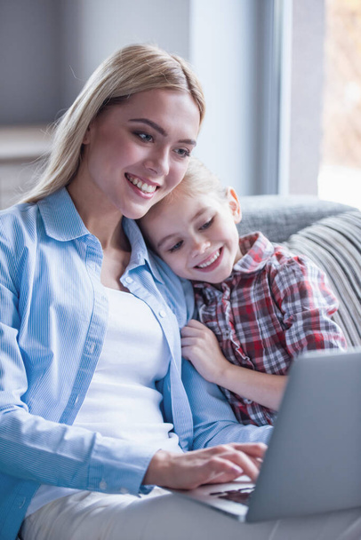 Mooie jonge moeder en haar schattig dochtertje zijn met behulp van een laptop en glimlachen terwijl het besteden van tijd samen - Foto, afbeelding