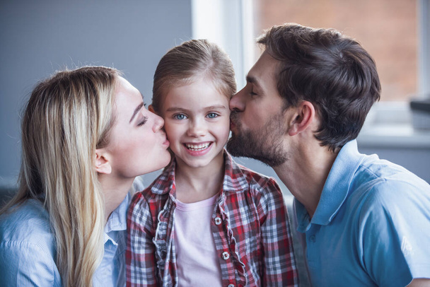 Beautiful young parents are kissing their cute little daughter, who is looking at camera and smiling - Photo, Image