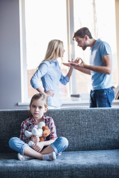 kleines Mädchen hält ein Spielzeug in der Hand und blickt traurig in die Kamera, während sich ihre Eltern im Hintergrund streiten - Foto, Bild