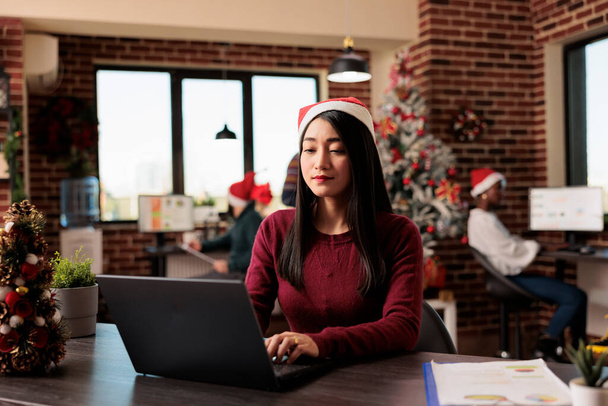 Азіатська жінка в капелюсі Санта керує кінцевим терміном проекту на ноутбуці, працюючи в декоративному офісі на Різдво. Співробітник компанії використовує комп'ютер на робочому місці під час зимового святкування сезону - Фото, зображення