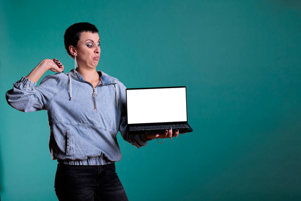 Angry unhappy woman with disapproval expression presenting laptop computer with blank screen for copyspace in studio. Sad negative female doing dislike gesture standing over isolated background. - Photo, Image
