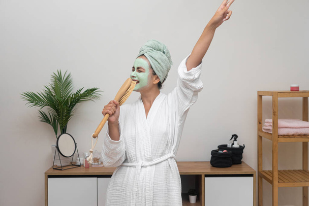 Femme heureuse avec masque d'argile sur le visage chantant en brosse à cheveux micro et dansant à la maison dame en peignoir avec serviette sur la tête s'amuser sur le soin de la peau de jour choyer - Photo, image