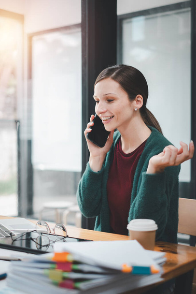 Привлекательная молодая женщина разговаривает по мобильному телефону и улыбается, сидя на своем рабочем месте в офисе и глядя на ноутбук
 - Фото, изображение