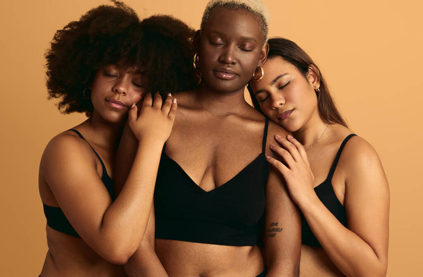 Tranquil young plus size multiethnic female models with long dark hair in black lingerie embracing African American friend with closed eyes against beige background - Foto, Bild