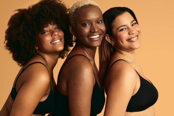Young happy diverse girlfriends in black lingerie with different skin color standing close and smiling against orange background looking at camera - Foto, Imagem