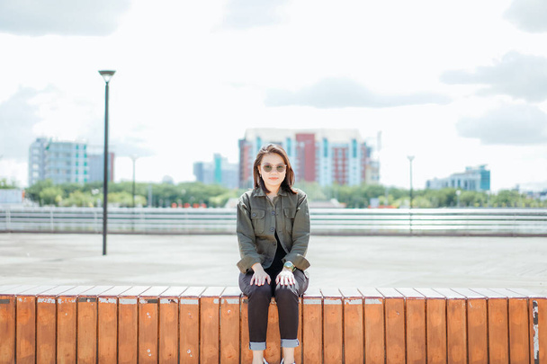 Молода красива азіатка одягнена куртка і чорні джинси позує на відкритому повітрі сидячи в парку - Фото, зображення