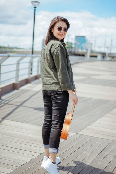 Играя Ukulele молодой красивой азиатской женщины носить куртку и черные джинсы Позирование на открытом воздухе - Фото, изображение