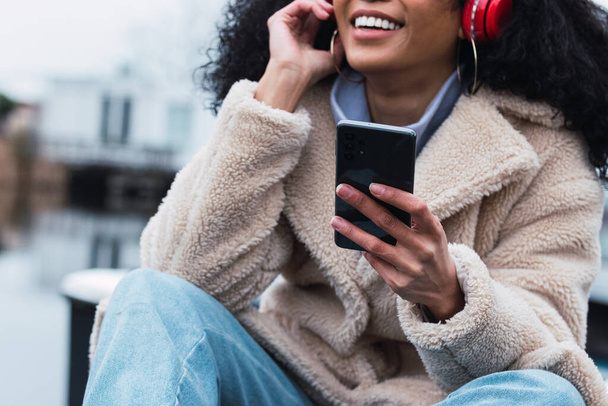 Mutlu İspanyol kadın cep telefonu kullanıyor, kulaklık takıyor ve açık havada müzik dinliyor. - Fotoğraf, Görsel