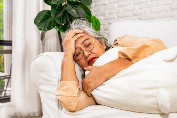 Elderly Asian woman sleep on sofa with frail health lying ill with ischemic stroke, hands touching temples headache chronic dizziness severe pain suffering from high blood pressure, cumulative stress. - Photo, Image